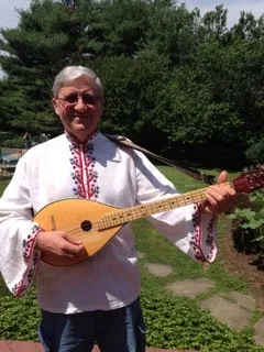 A gentleman in traditional Bulgarian embroidered white top with red and black designs. He is holding a Bulgarian tambura (long necked string instrument).